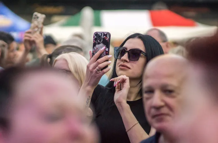 Eine Frau mit Sonnenbrille hält ihr Smartphone hoch, um in einer Menschenmenge ein Foto oder Video zu machen.