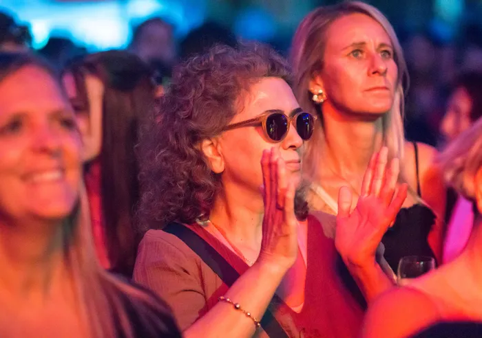 Eine Frau mit lockigem Haar und Sonnenbrille hebt die Hände bei einer Abendveranstaltung unter warmer Beleuchtung.