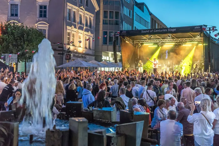 Eine große Menschenmenge feiert vor einer hell erleuchteten Bühne bei einem Open-Air-Konzert in einer Stadt.