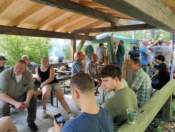 Eine Gruppe von Männern sitzt und steht unter einem Holzpavillon zusammen, einige von ihnen schauen auf ein Smartphone oder unterhalten sich. Im Hintergrund ist ein Grill zu sehen, über dem Rauch aufsteigt.