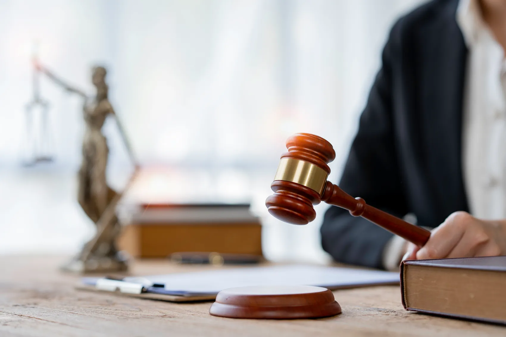 Ein Richter oder eine Richterin schlägt mit einem hölzernen Hammer auf eine Schallplatte. Im Hintergrund sind eine Justizstatue und Bücher zu sehen.