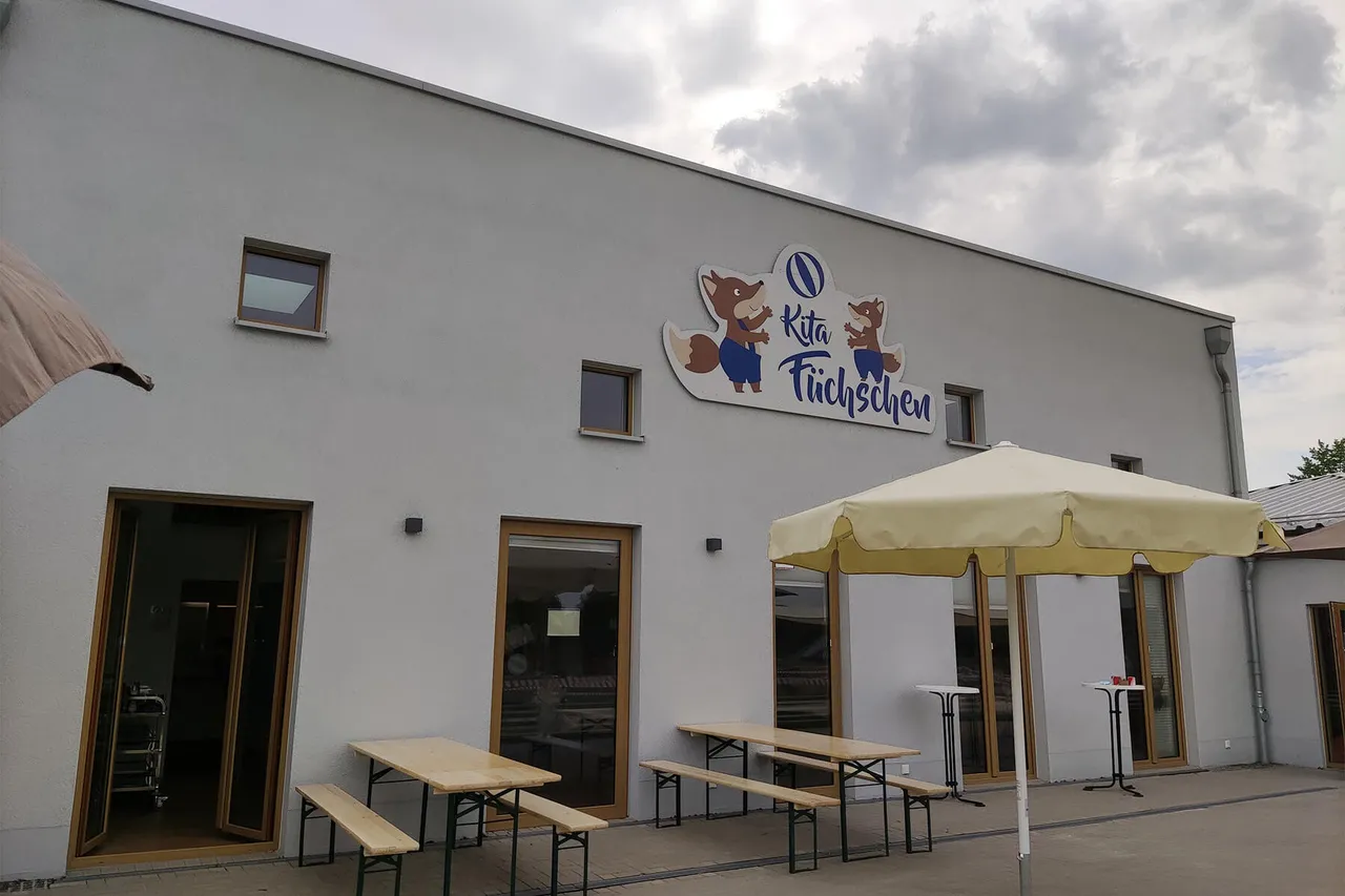 Ein hellgraues Gebäude mit dem Schild „Kita Füchschen“ über dem Eingangsbereich. Vor dem Gebäude stehen Tische mit Sonnenschirmen auf einem gepflasterten Vorplatz.