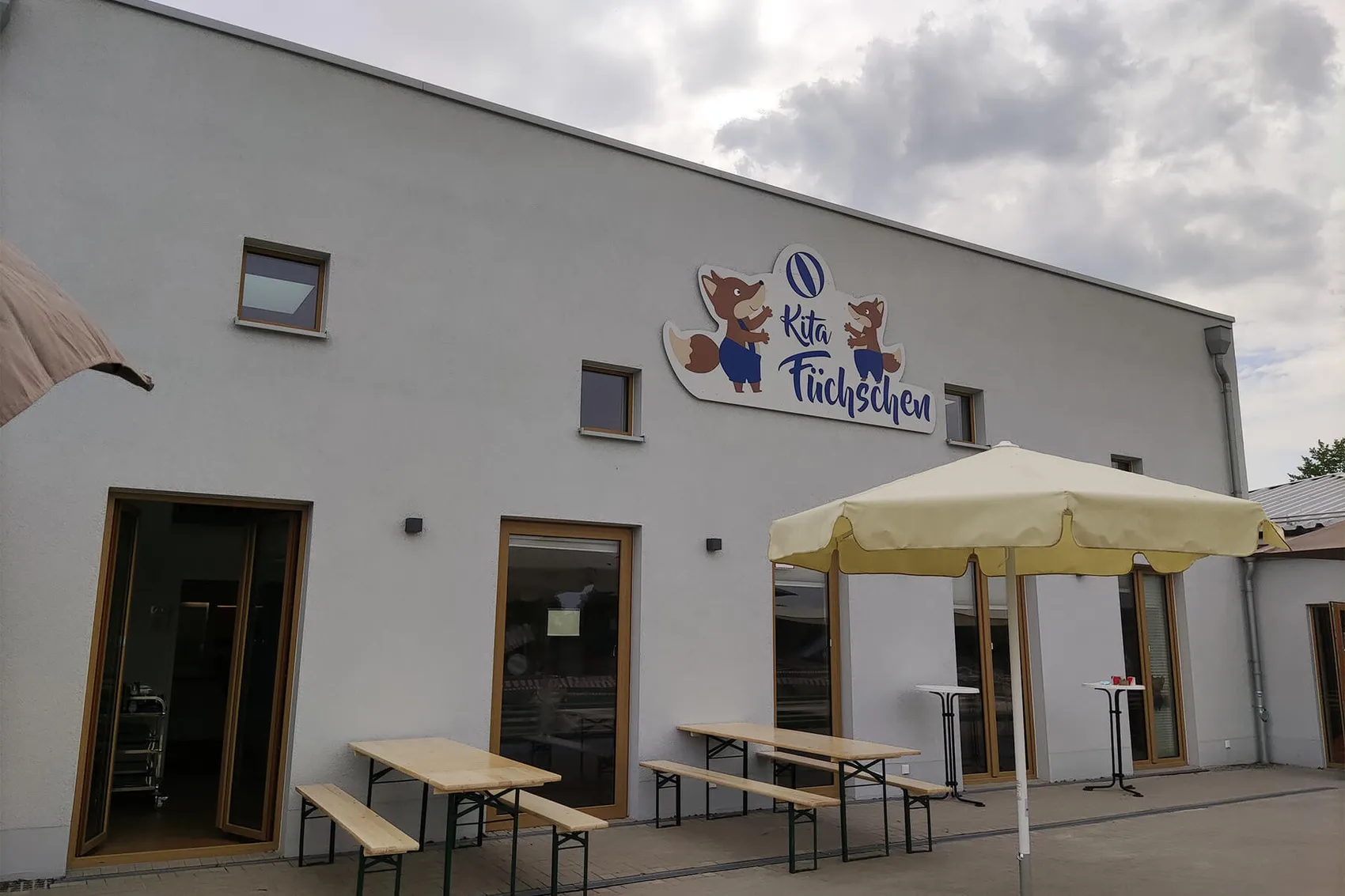 Ein hellgraues Gebäude mit dem Schild „Kita Füchschen“ über dem Eingangsbereich. Vor dem Gebäude stehen Tische mit Sonnenschirmen auf einem gepflasterten Vorplatz.