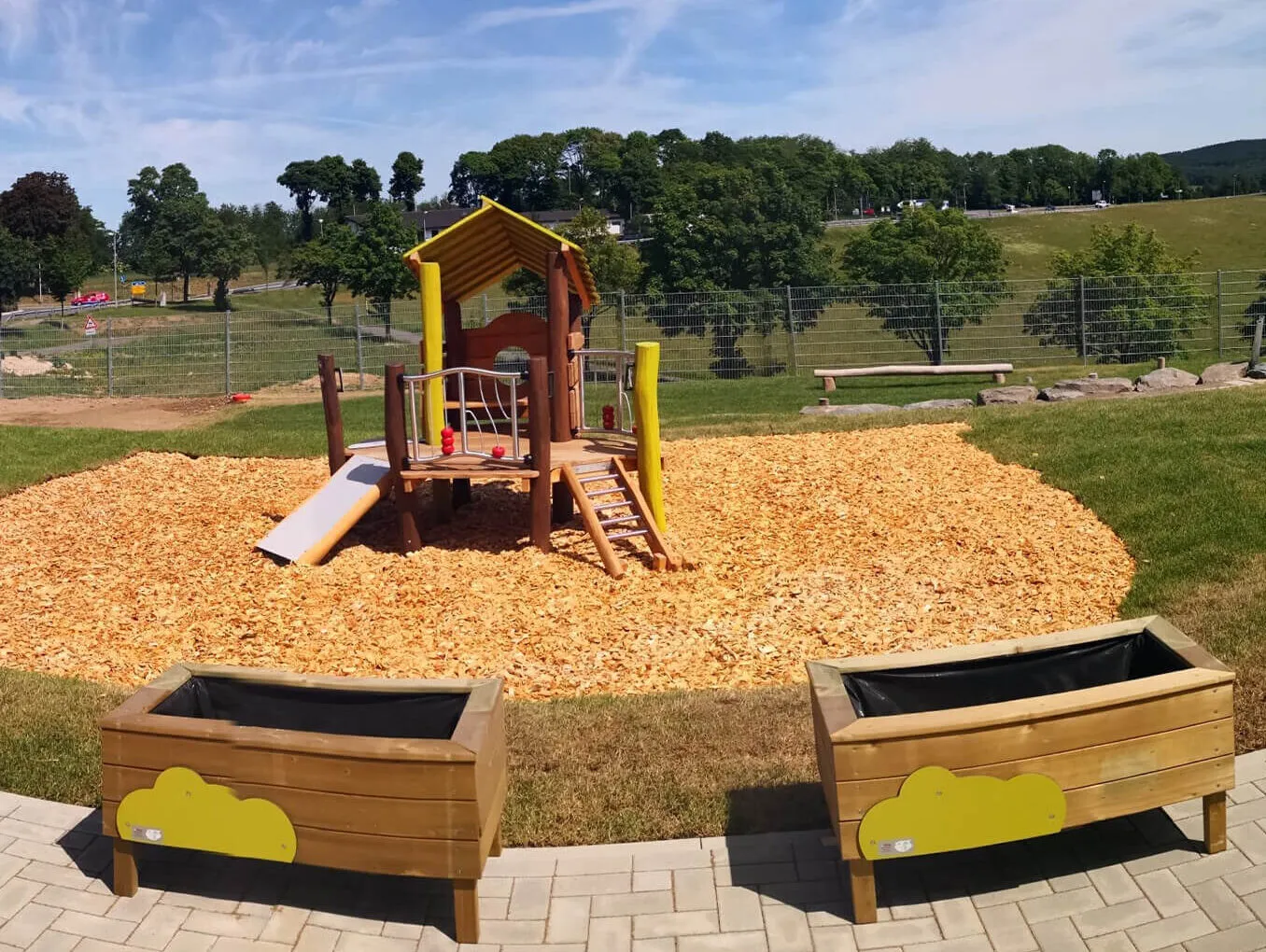 Spielturm mit Rutsche und Klettergerüst auf einem Spielplatz mit Holzschnitzelboden. Im Vordergrund zwei Sandkästen mit gelben Wolkenmotiven.