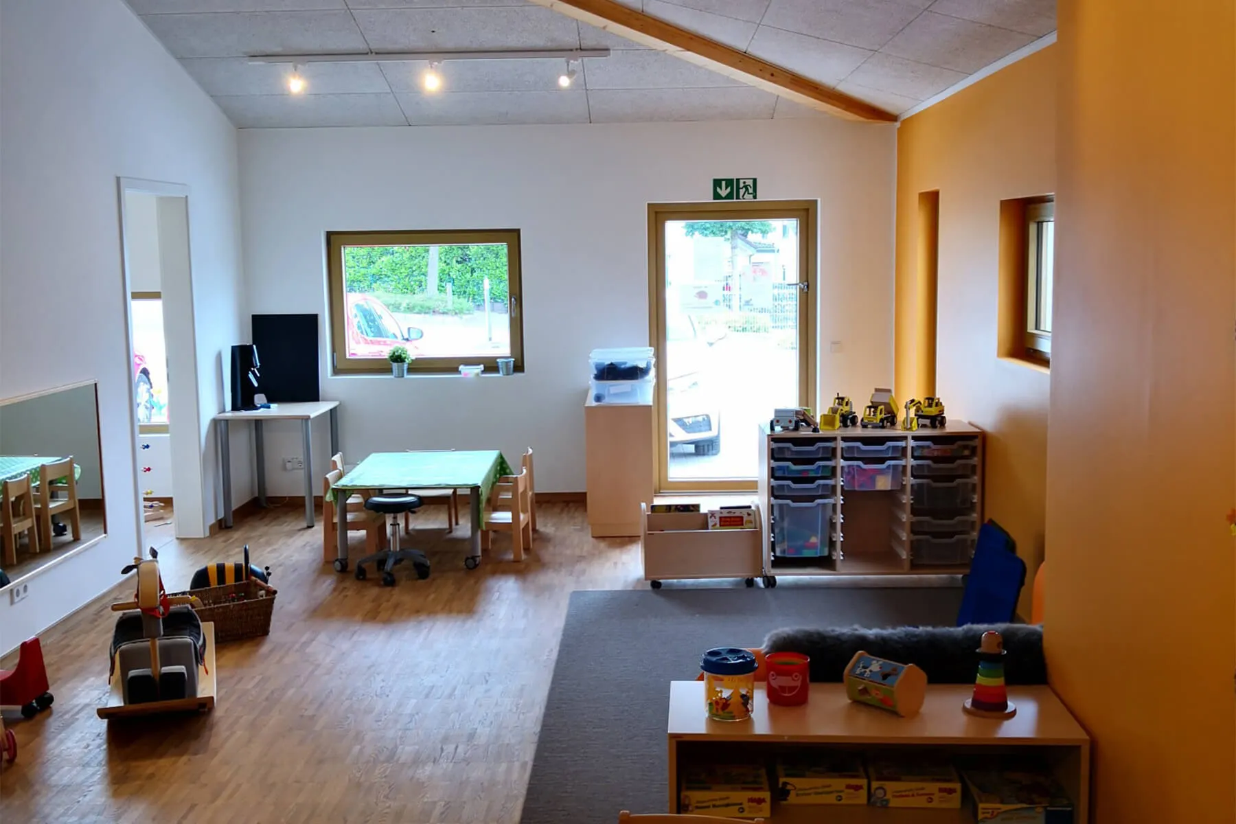 Heller Gruppenraum einer Kindertagesstätte mit Spieltischen, Spielzeug und einer offenen Tür zu einem Außenbereich.