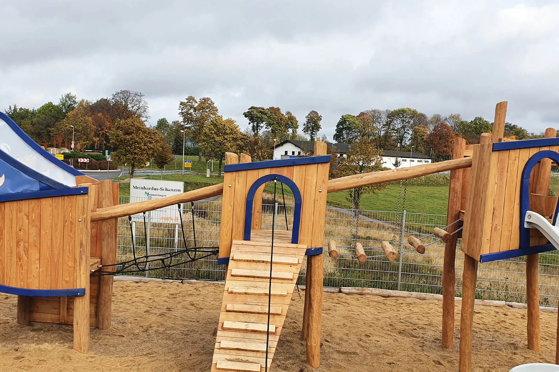 Holzspielplatz mit Klettergerüsten, Rutschen und Seilen vor einer Hügellandschaft und Häusern.