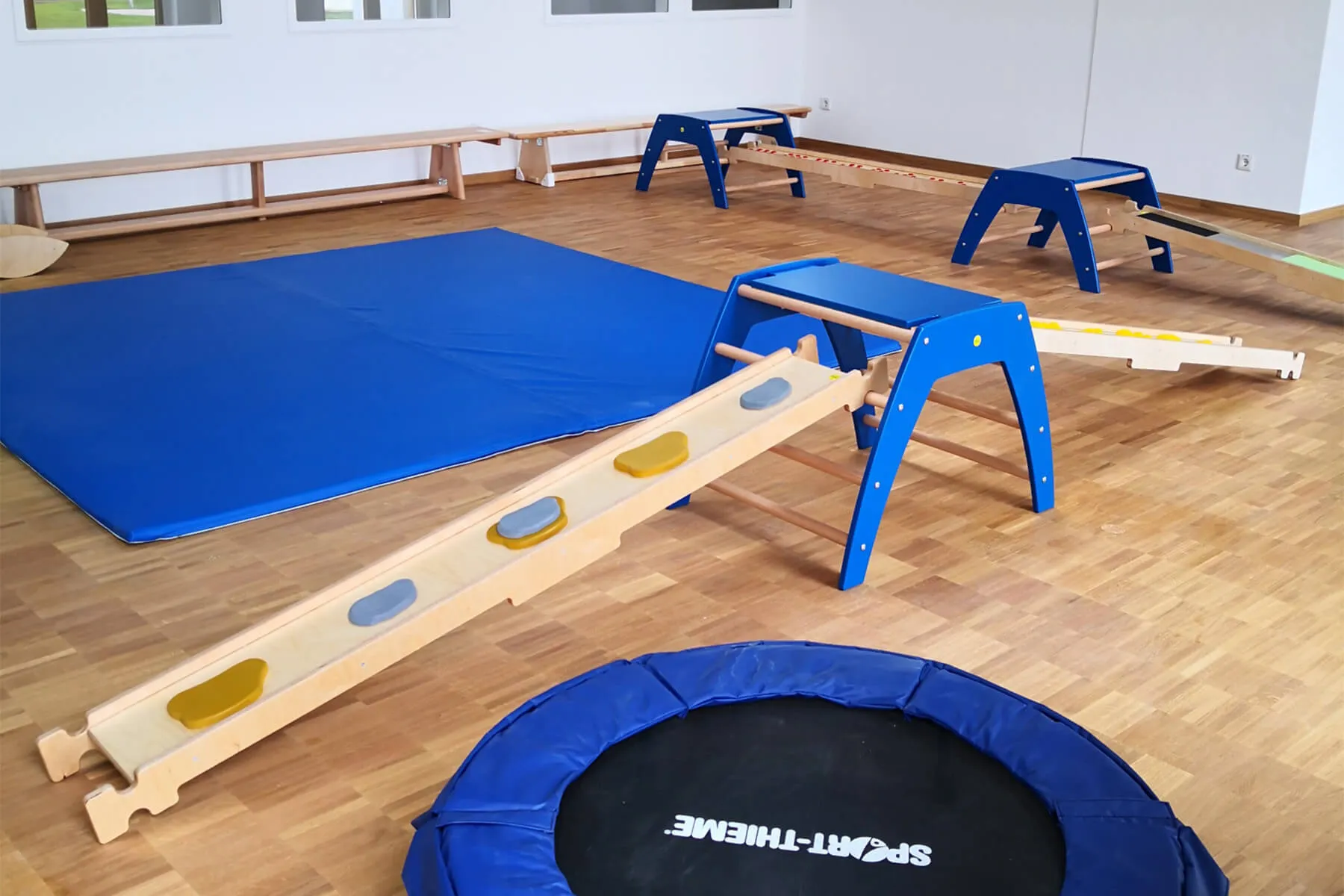 Ein heller Mehrzweckraum in einer Kita mit blauen Matten, einem Trampolin und verschiedenen Holzgeräten für Kinder zum Klettern und Balancieren.