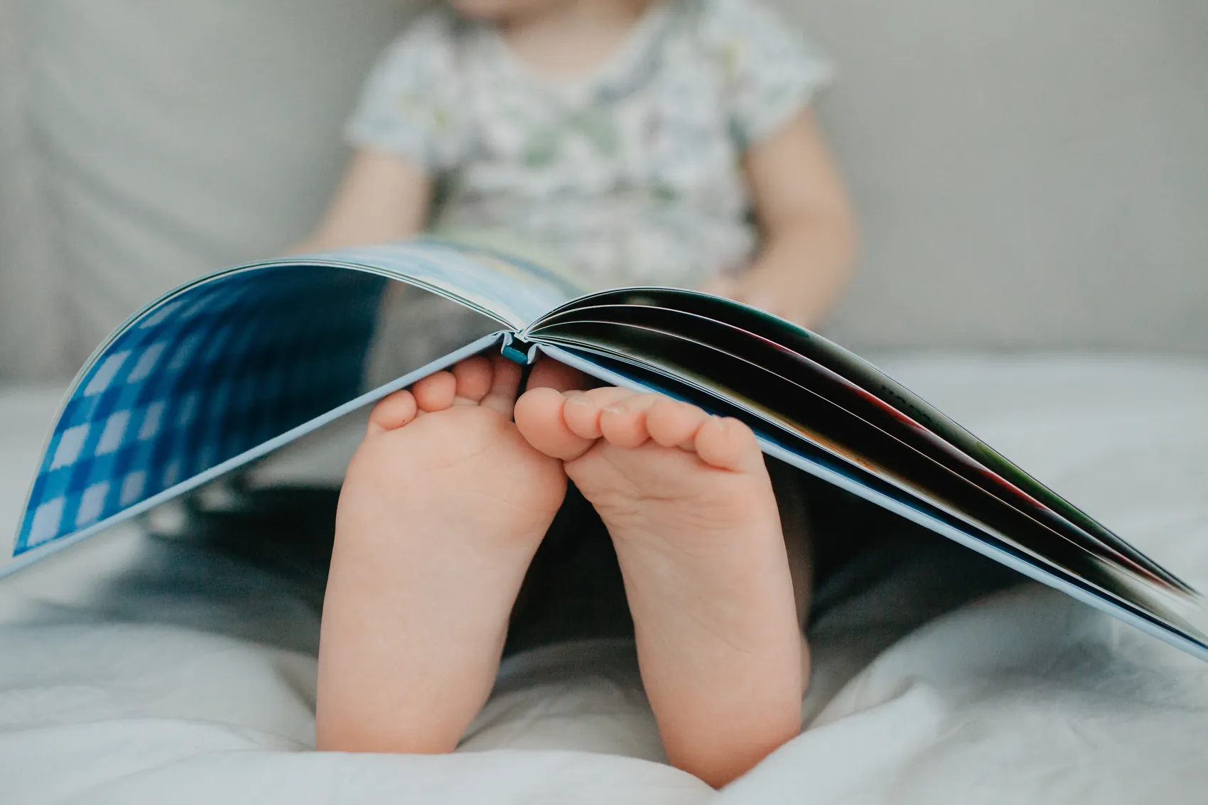 Ein Kind liest ein aufgeschlagenes Buch. Im Fokus stehen die nackten Füße des Kindes, während der Rest des Bildes unscharf ist.