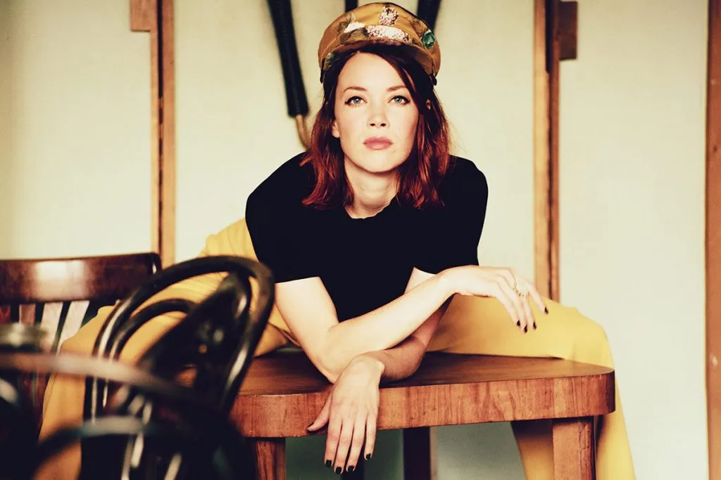 Magdalena Ganter, rothaarige Sängerin mit einer Schirmmütze, lehnt an einem Holztisch im Kulturhaus.