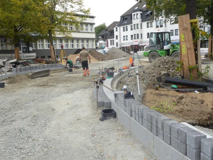 Bauarbeiten auf dem Maiplatz mit Arbeitern, einem grünen Radlader und neu gesetzten Bordsteinen vor Wohnhäusern.