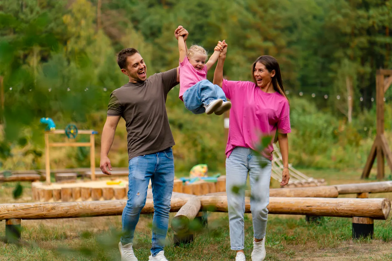 Ein lächelndes Paar geht mit seiner kleinen Tochter auf einem Waldweg spazieren, das Mädchen wird in die Höhe geworfen.