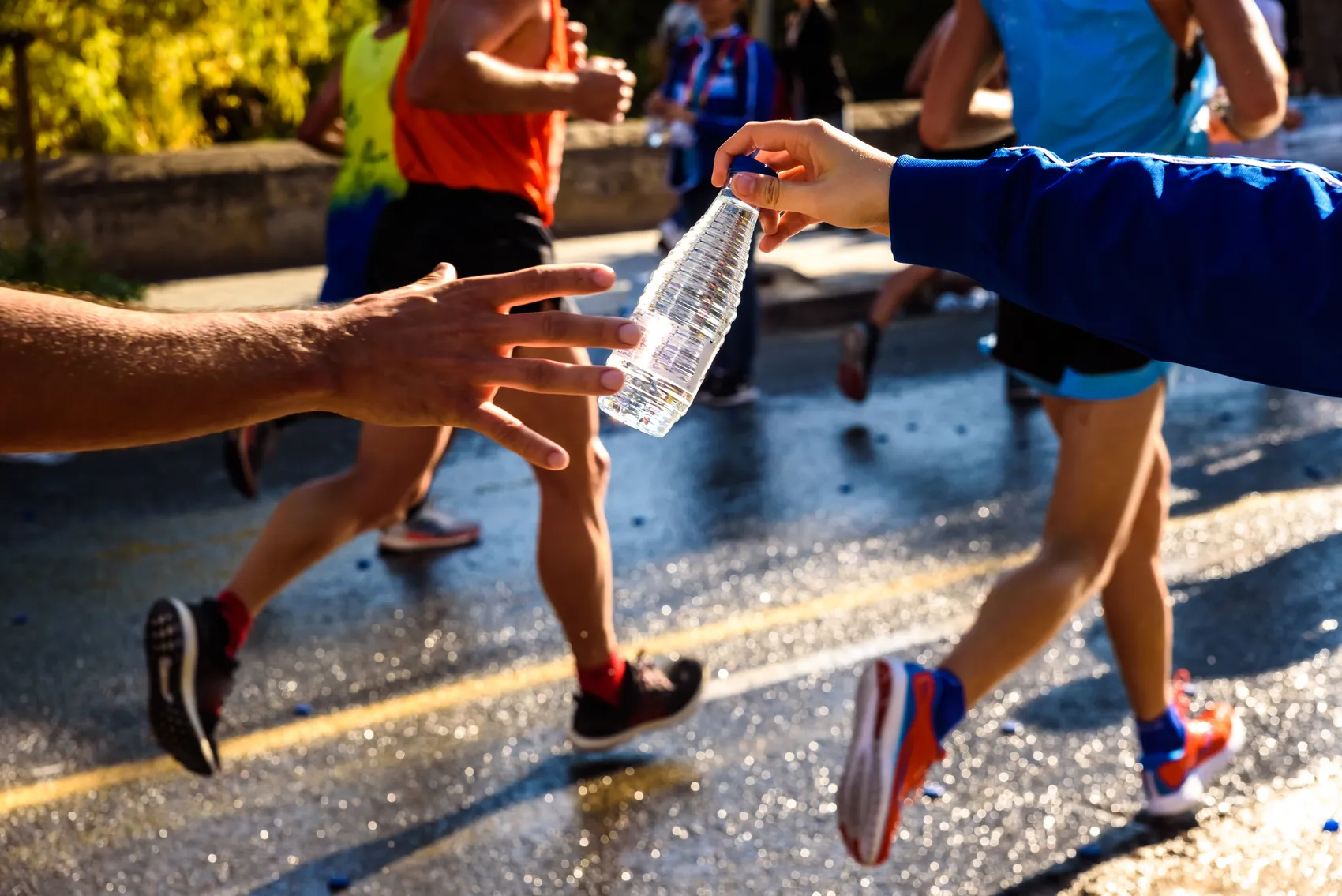 Ein Läufer nimmt während eines Marathons eine Trinkflasche von einer Person am Straßenrand entgegen. Die Straße ist nass und mehrere andere Läufer sind im Hintergrund zu sehen.