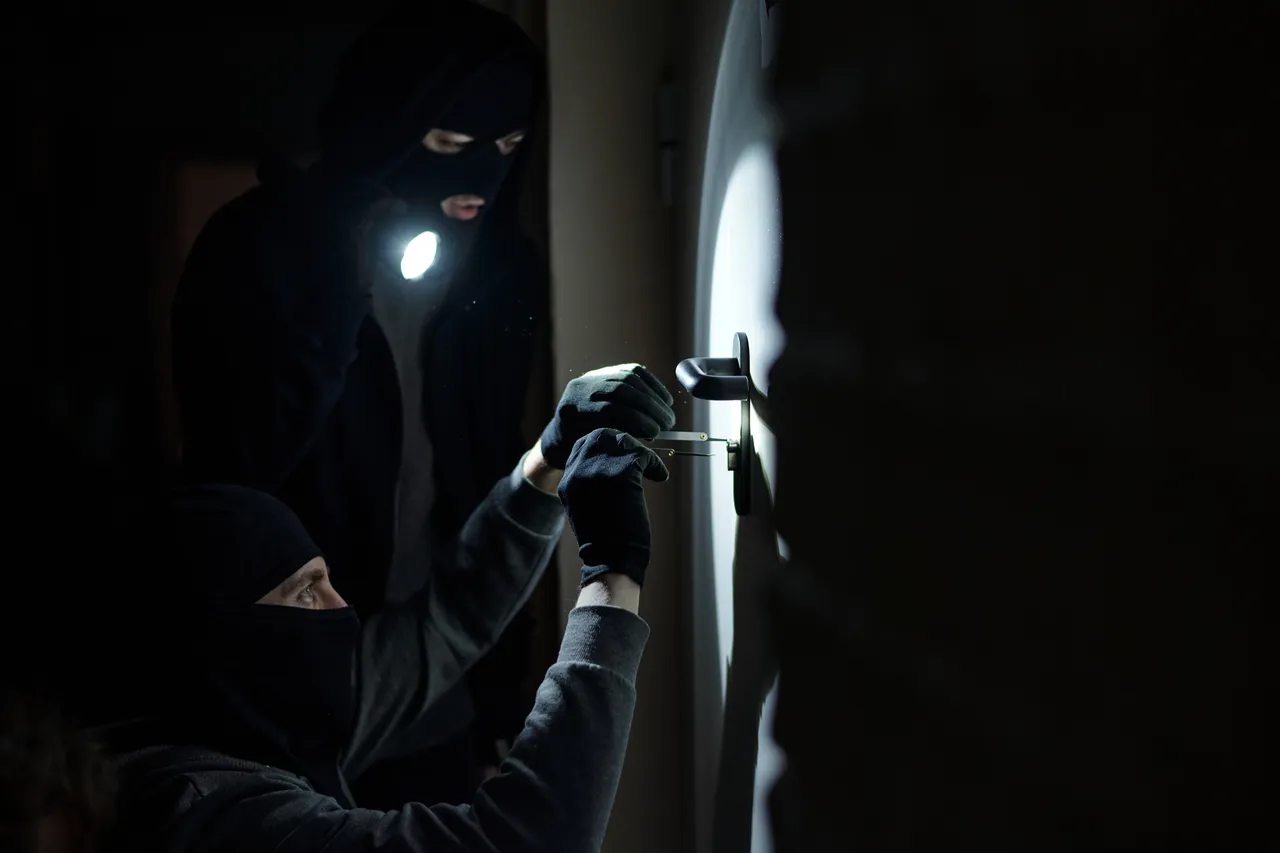 Zwei maskierte Personen versuchen bei Dunkelheit, ein Türschloss mit einem Werkzeug zu öffnen, während eine Person mit einer Taschenlampe leuchtet.