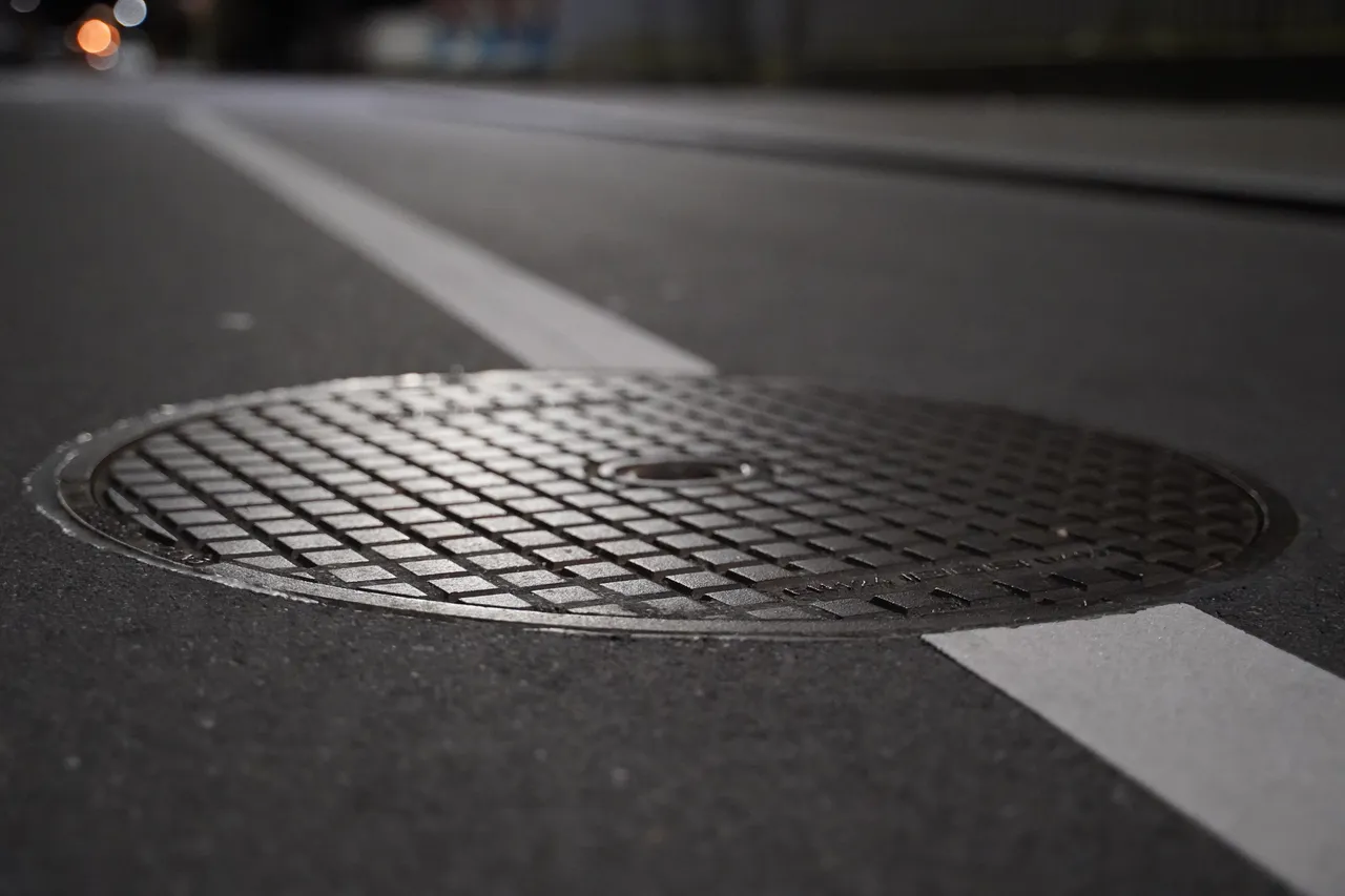 Nahaufnahme eines runden Metall-Gullydeckels auf einer asphaltierten Straße mit einer weißen Markierungslinie.