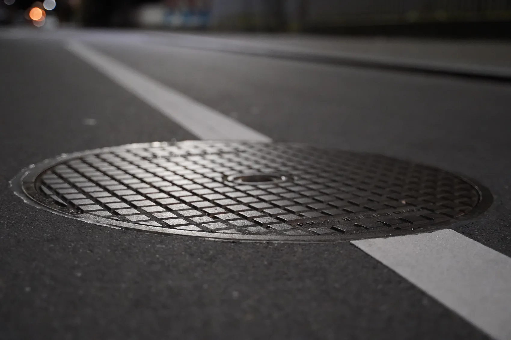 Nahaufnahme eines runden Metall-Gullydeckels auf einer asphaltierten Straße mit einer weißen Markierungslinie.