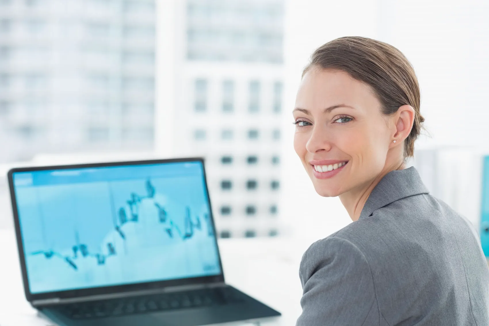 Lachende Frau im Business-Kostüm blickt über die Schulter in die Kamera, während auf einem Laptop vor ihr ein Diagramm mit steigenden und fallenden Linien zu sehen ist.