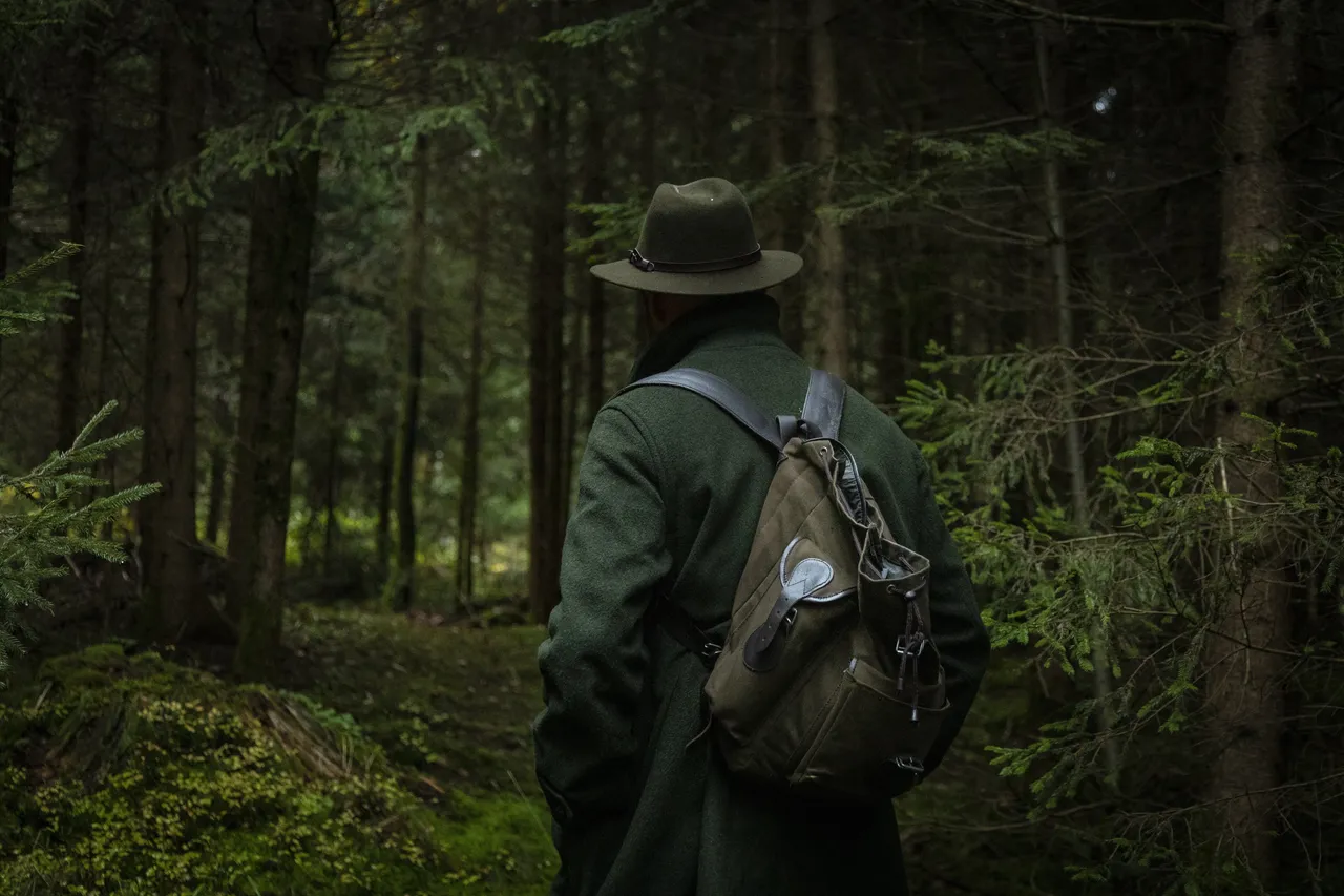 Ein Mann mit Hut, grünem Mantel und Rucksack steht mit dem Rücken zum Betrachter in einem dichten Wald.