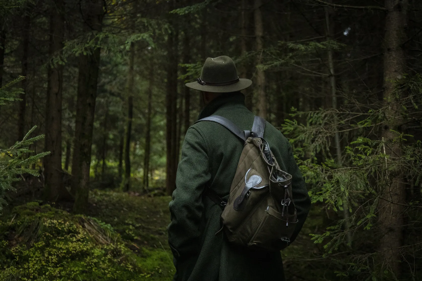 Ein Mann mit Hut, grünem Mantel und Rucksack steht mit dem Rücken zum Betrachter in einem dichten Wald.