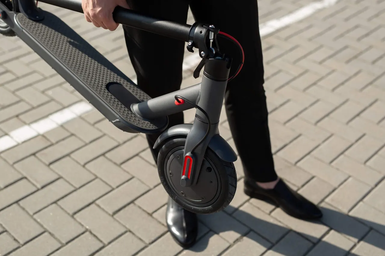 Eine Person in schwarzer Kleidung hält einen schwarzen E-Scooter über einem gepflasterten Boden.