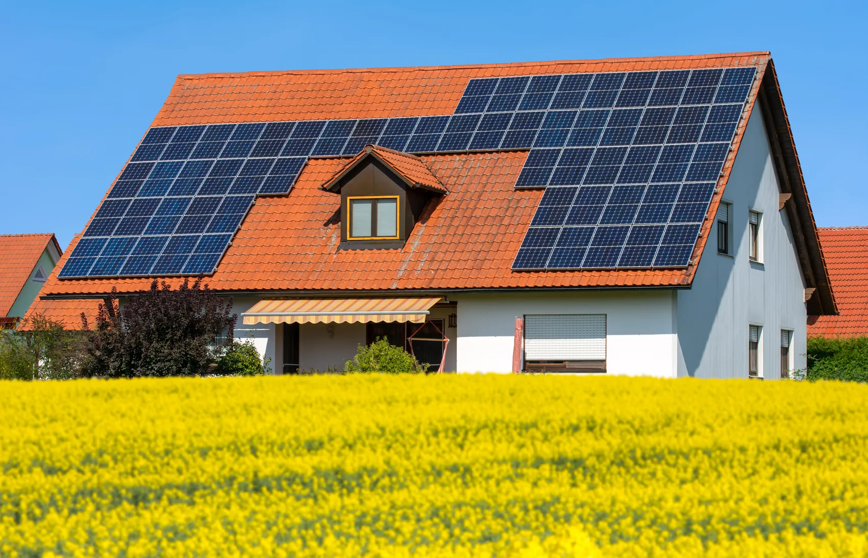 Ein Haus mit roten Ziegeln und einem großen Solarmodul auf dem Dach steht vor einem Feld mit gelben Blüten.