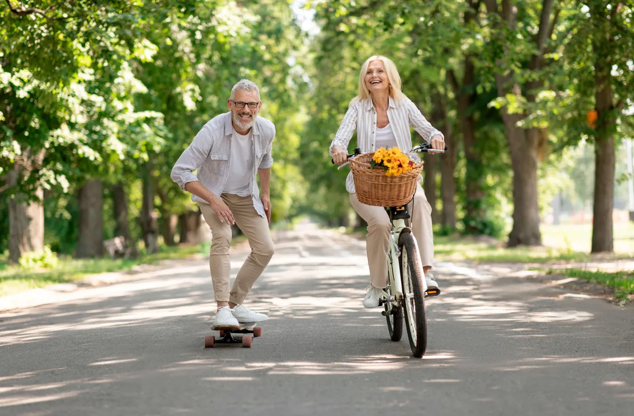 Ein älteres Paar genießt einen sonnigen Tag auf einer Landstraße: Die Frau fährt Fahrrad mit einem Blumenkorb, der Mann fährt auf einem Skateboard.