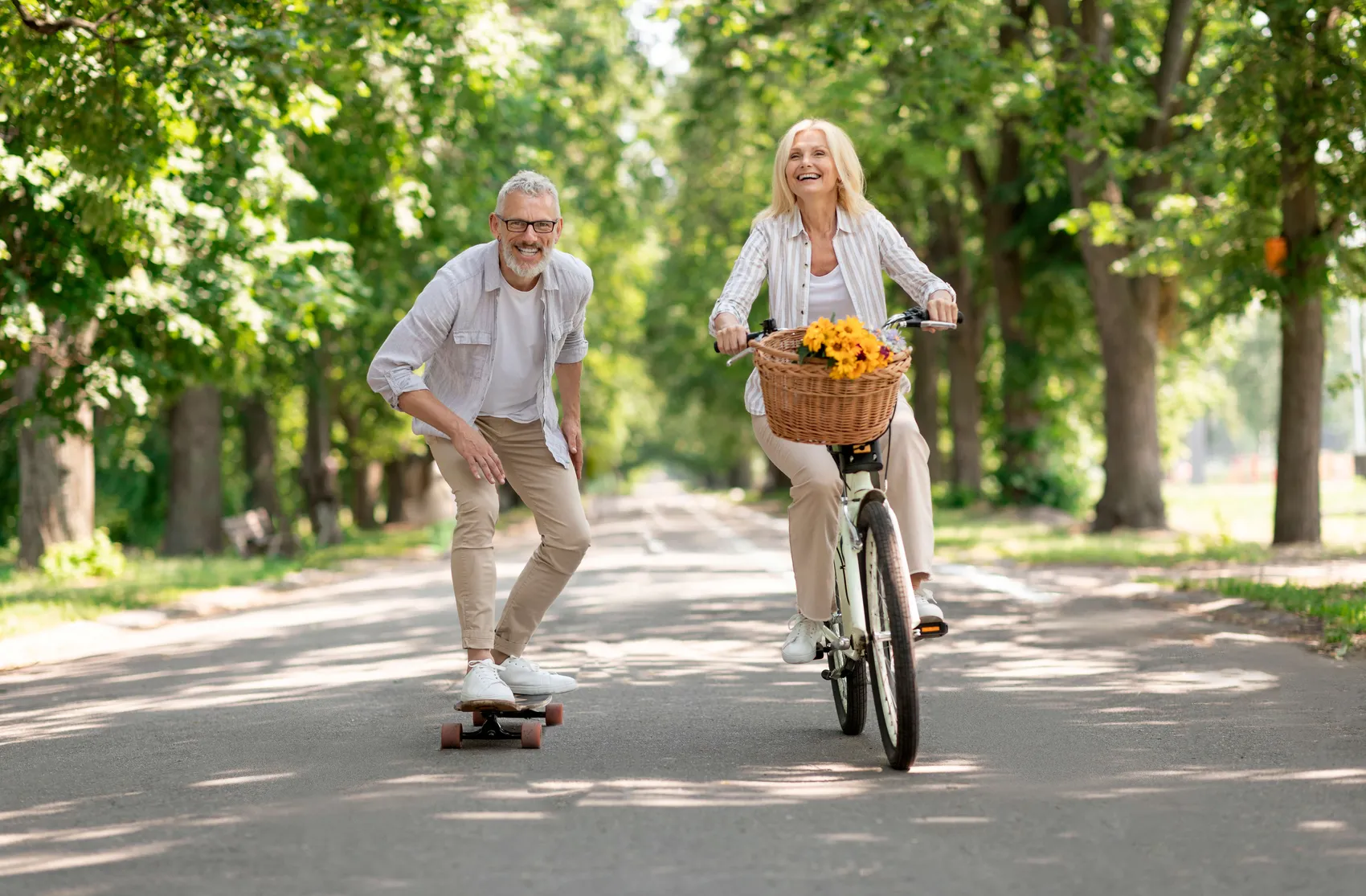 Ein älteres Paar genießt einen sonnigen Tag auf einer Landstraße: Die Frau fährt Fahrrad mit einem Blumenkorb, der Mann fährt auf einem Skateboard.