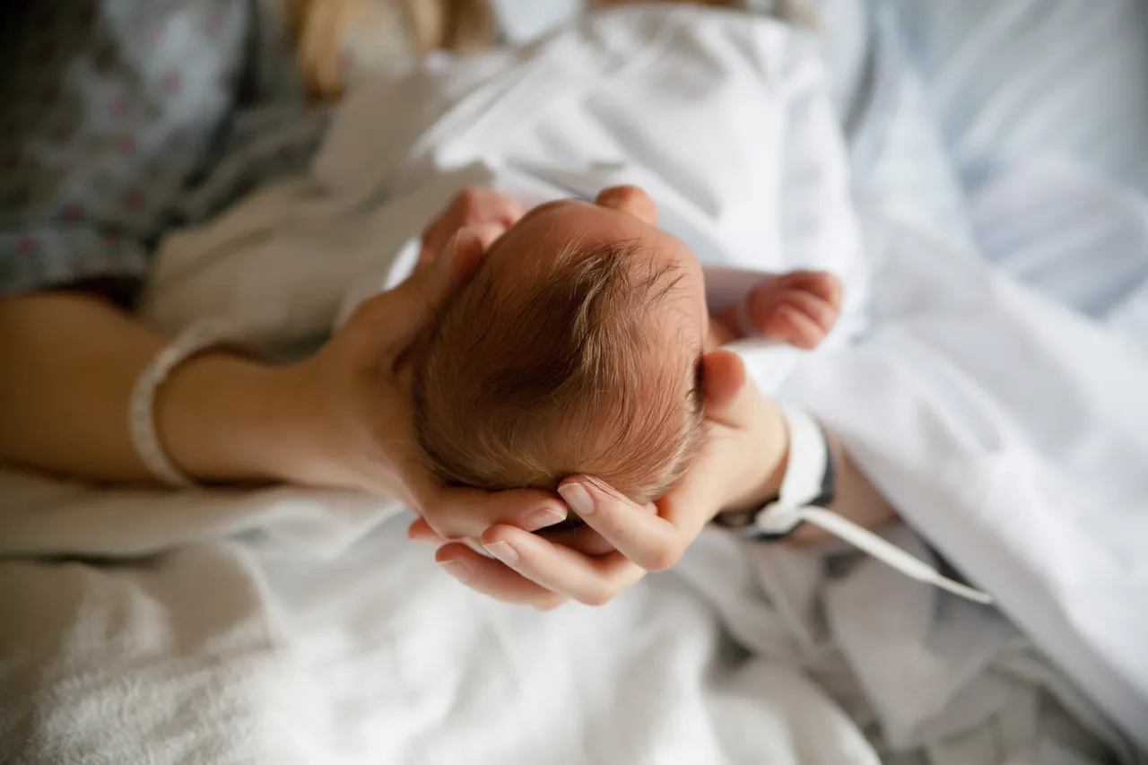 Eine Mutter hält das Köpfchen ihres Neugeborenen in ihren Händen. Beide tragen Armbänder, das Baby liegt auf einer weißen Bettdecke.