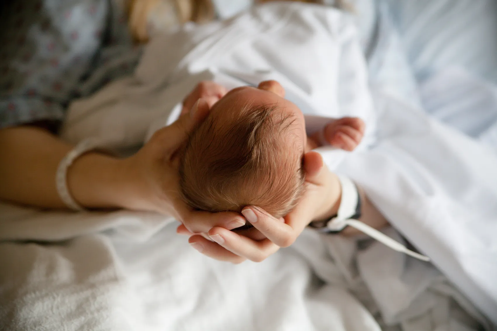 Eine Mutter hält das Köpfchen ihres Neugeborenen in ihren Händen. Beide tragen Armbänder, das Baby liegt auf einer weißen Bettdecke.