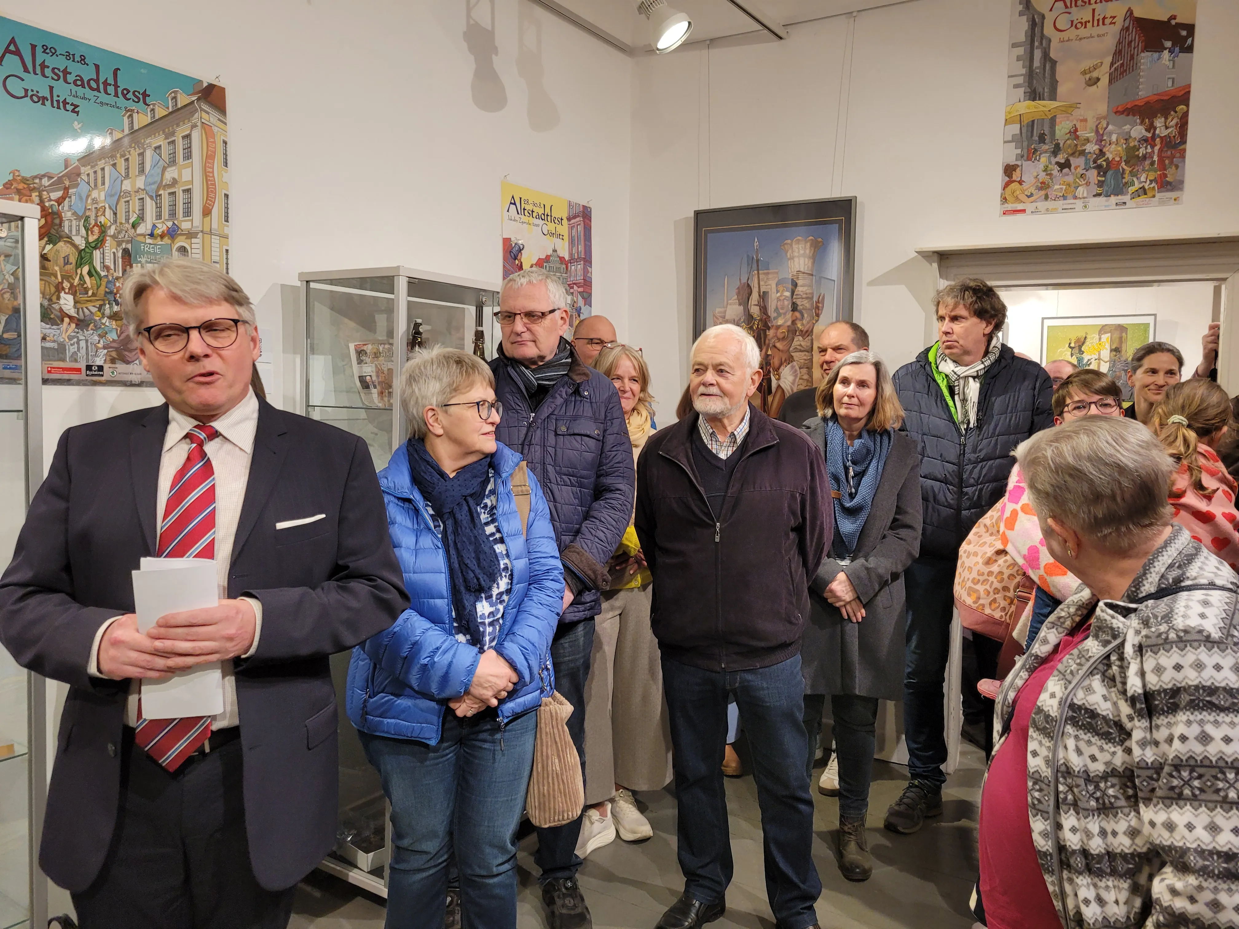 Mann mit Mikrofon spricht vor einer Gruppe von Menschen in einem Ausstellungsraum, im Hintergrund Gemälde und Plakate mit Stadtansichten.