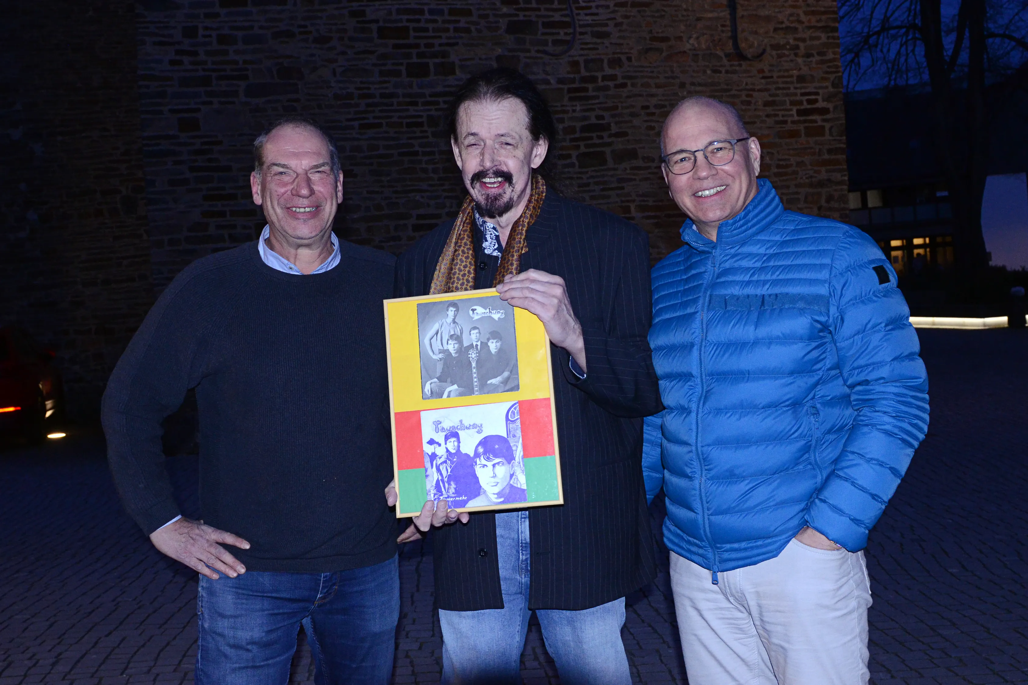 Drei Männer posieren vor einer Backsteinmauer. Der Mann in der Mitte hält ein Buch mit zwei Covern, darunter ein frühes Werk der Band „Täuschung“.