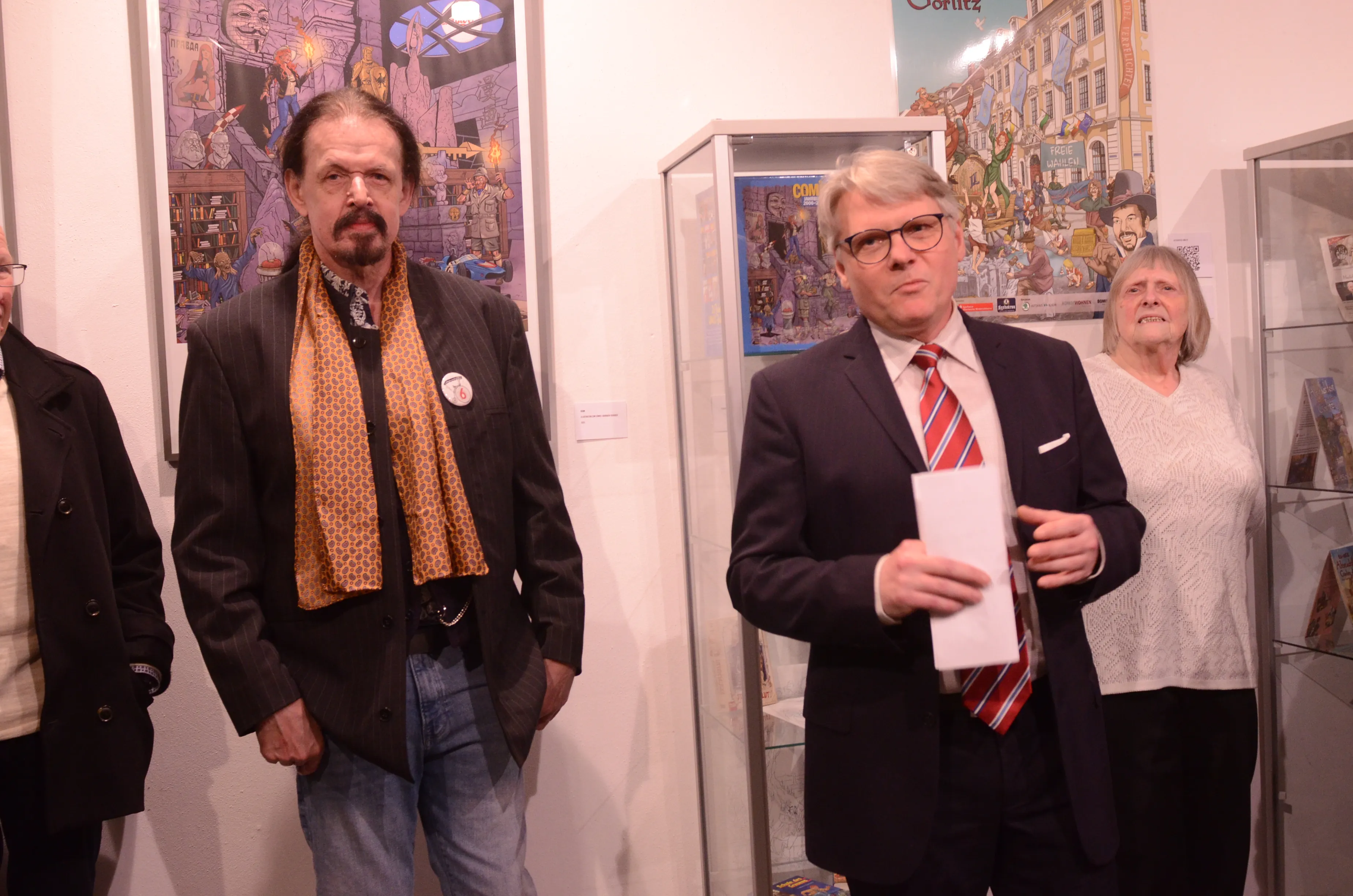 Drei Personen stehen vor Comiczeichnungen in einer Ausstellung. Ein Mann in Anzug und roter Krawatte hält Papier in der Hand, während eine Frau neben ihm spricht. Im Hintergrund sind weitere Kunstwerke und Vitrinen zu sehen.
