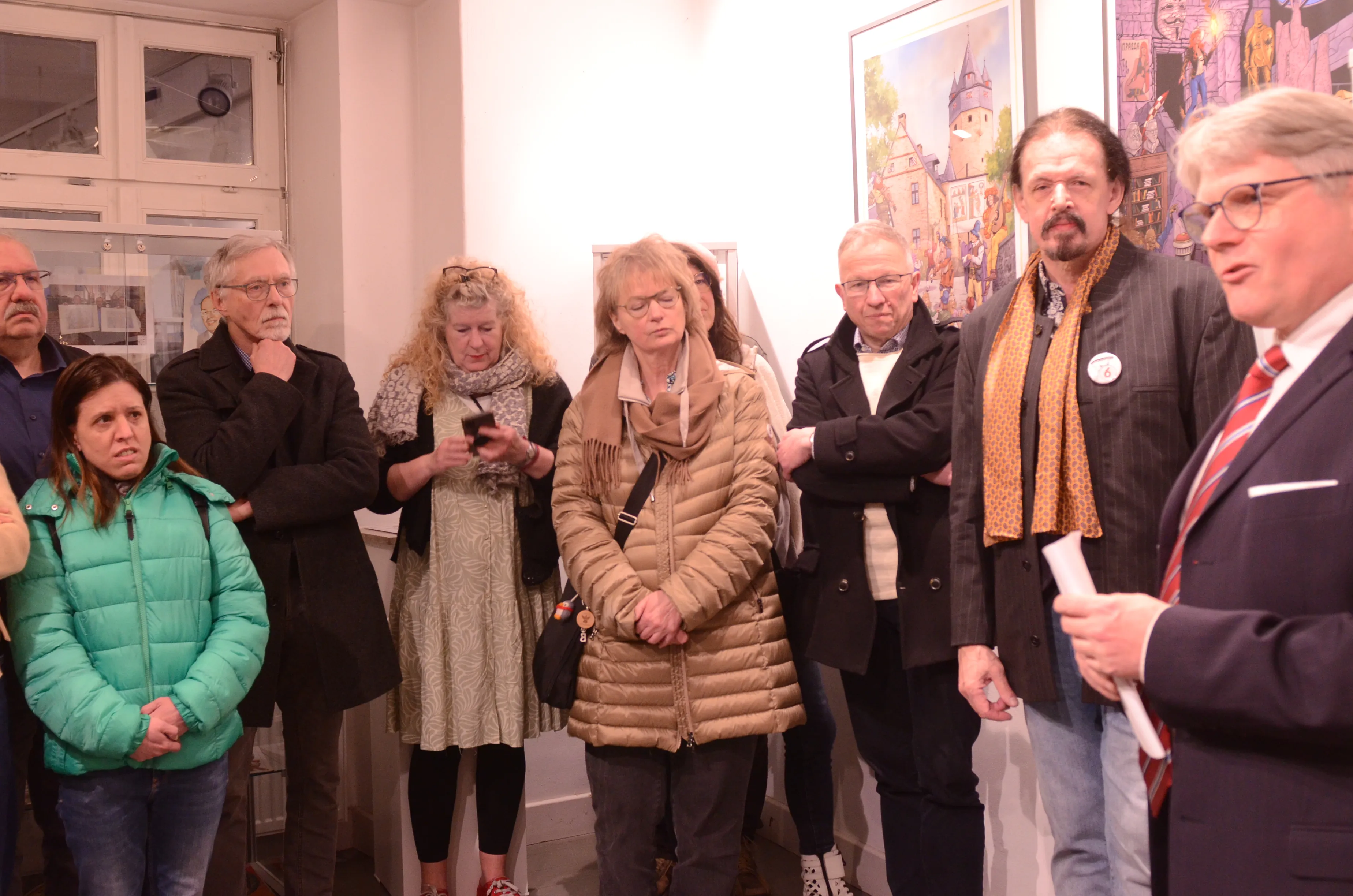 Eine Gruppe von Menschen steht in einem Raum vor Gemälden, während ein Mann mit Bart und Anzug spricht. Im Hintergrund sind weitere Kunstwerke an der Wand zu sehen.