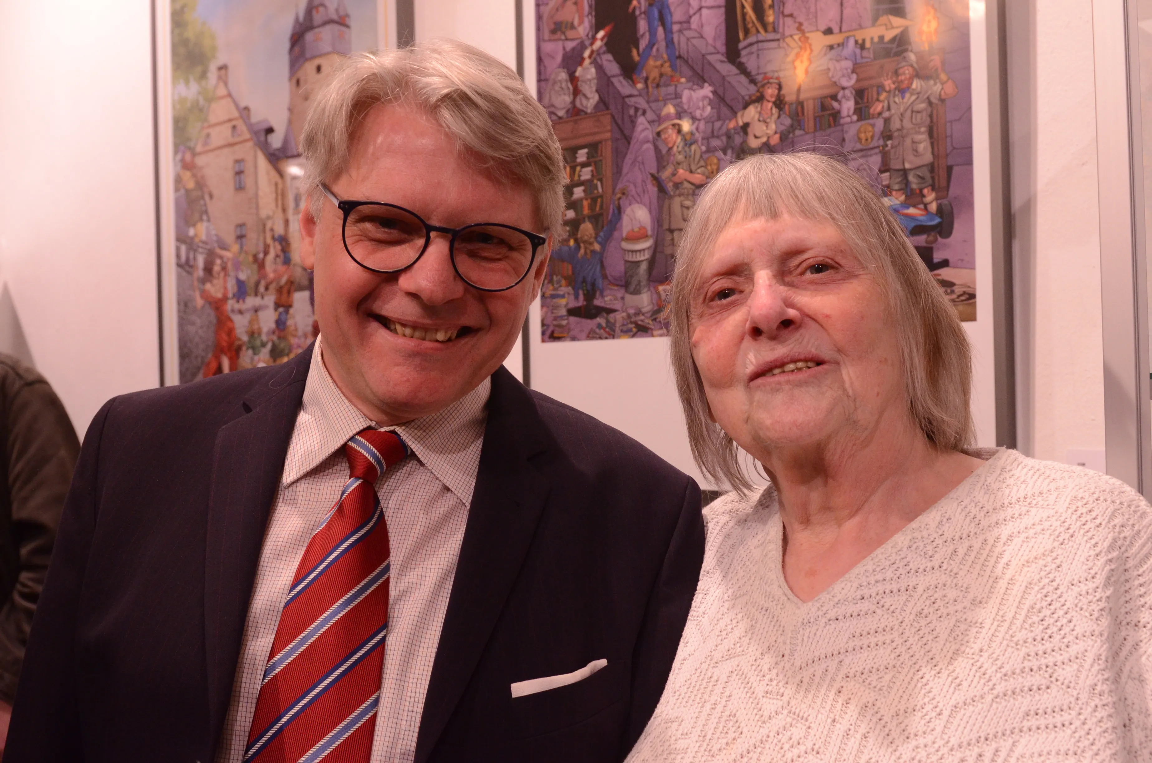 Dietmar Krüger, Comiczeichner und Illustrator, lächelt neben einer älteren Frau vor einem farbenfrohen Gemälde einer mittelalterlichen Stadtansicht.