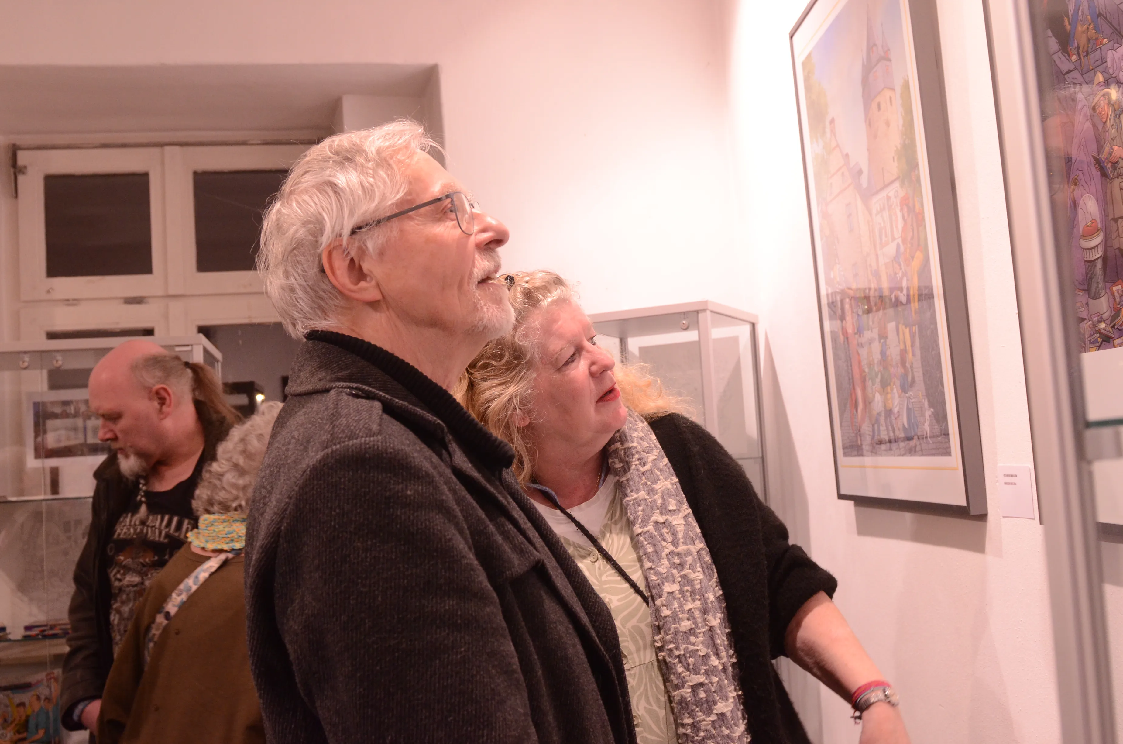 Zwei ältere Besucher betrachten ein farbenfrohes Gemälde einer Stadtansicht in einer Ausstellung im Heimathaus.