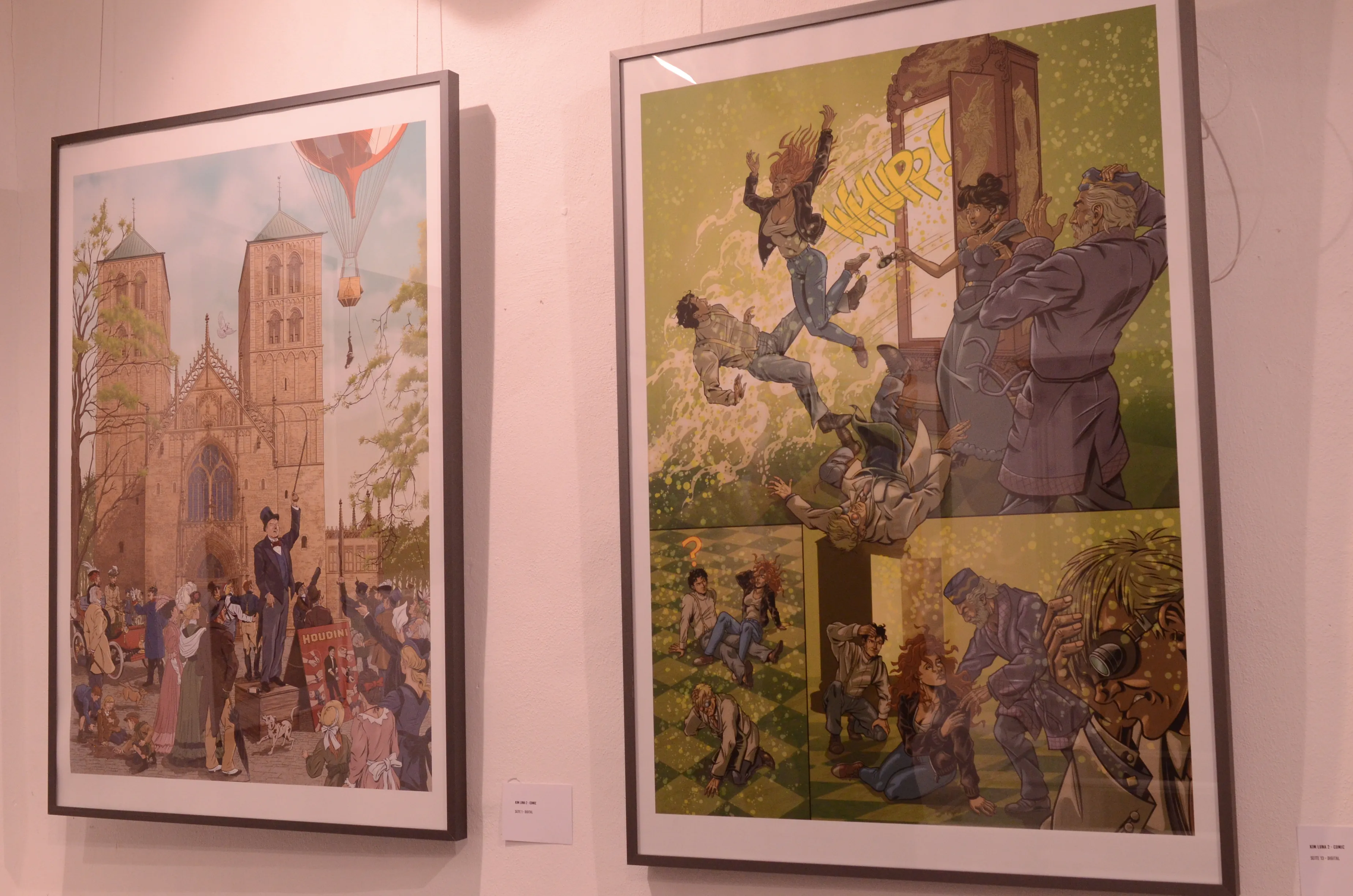Zwei gerahmte Comiczeichnungen hängen an einer weißen Wand. Die linke zeigt eine historische Szene mit einer Menschenmenge vor einer Kirche, während die rechte eine dynamische Szene mit fallenden Figuren und Trümmern darstellt.