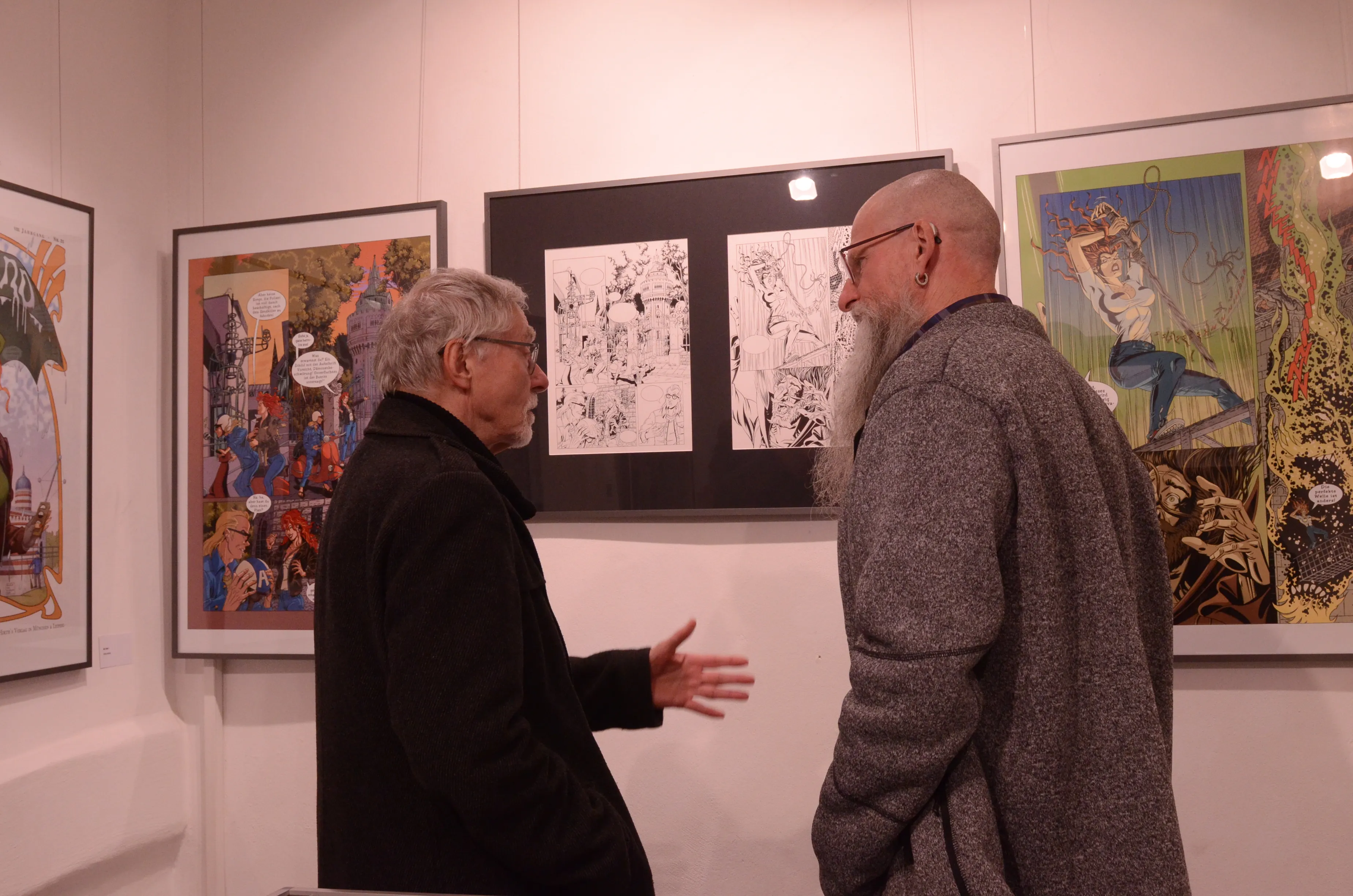 Zwei ältere Männer betrachten in einem hellen Raum Comiczeichnungen an den Wänden. Im Hintergrund sind weitere Bilder zu sehen.
