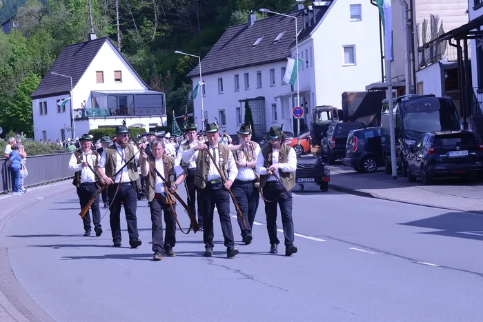 Eine Gruppe von Männern in traditioneller Tracht marschiert mit Gewehren auf einer Straße während eines Festumzugs. Im Hintergrund sind Häuser und Zuschauer zu sehen.