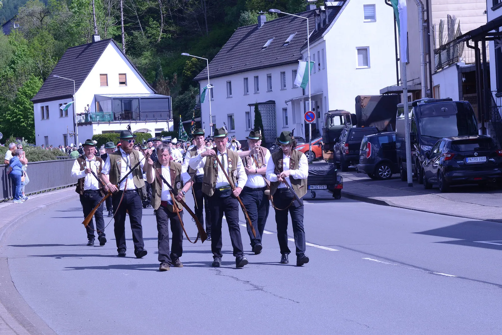 Eine Gruppe von Männern in traditioneller Tracht marschiert mit Gewehren auf einer Straße während eines Festumzugs. Im Hintergrund sind Häuser und Zuschauer zu sehen.