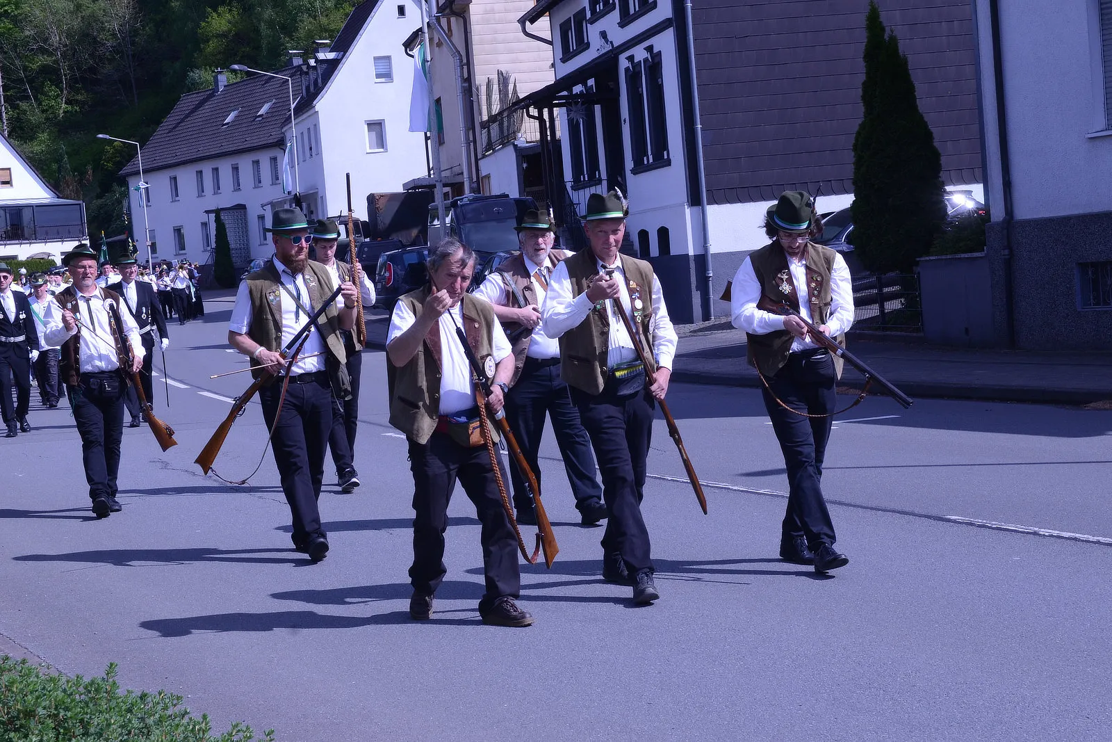Eine Gruppe von Männern in traditioneller Tracht mit Gewehren marschiert auf einer Straße während eines Festumzugs.