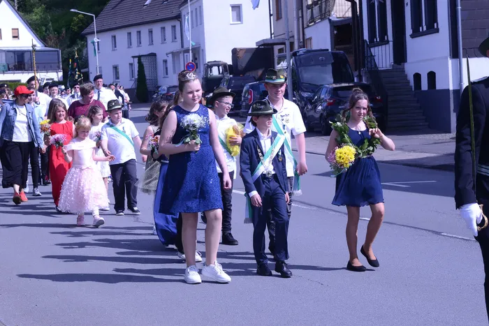 Festumzug mit Kindern und Erwachsenen in festlicher Kleidung, einige tragen Orden und Blumensträuße. Im Hintergrund sind Häuser und ein Fahrzeug zu sehen.