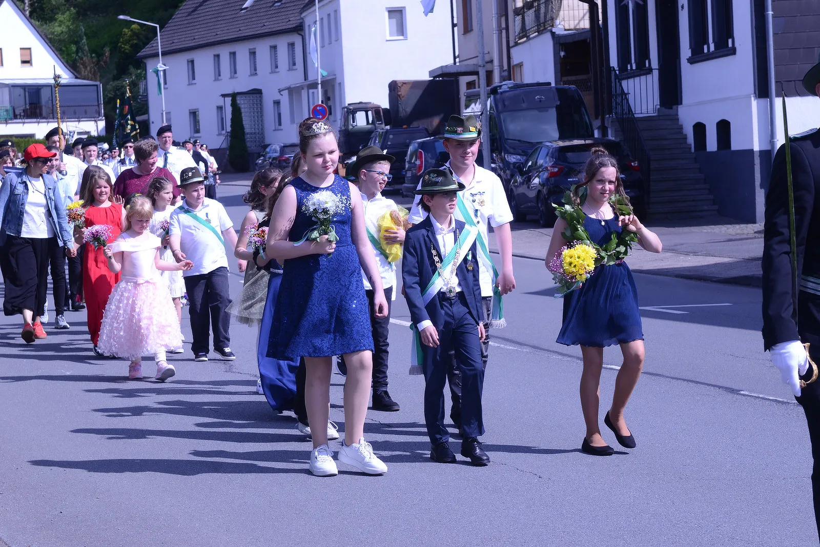 Festumzug mit Kindern und Erwachsenen in festlicher Kleidung, einige tragen Orden und Blumensträuße. Im Hintergrund sind Häuser und ein Fahrzeug zu sehen.