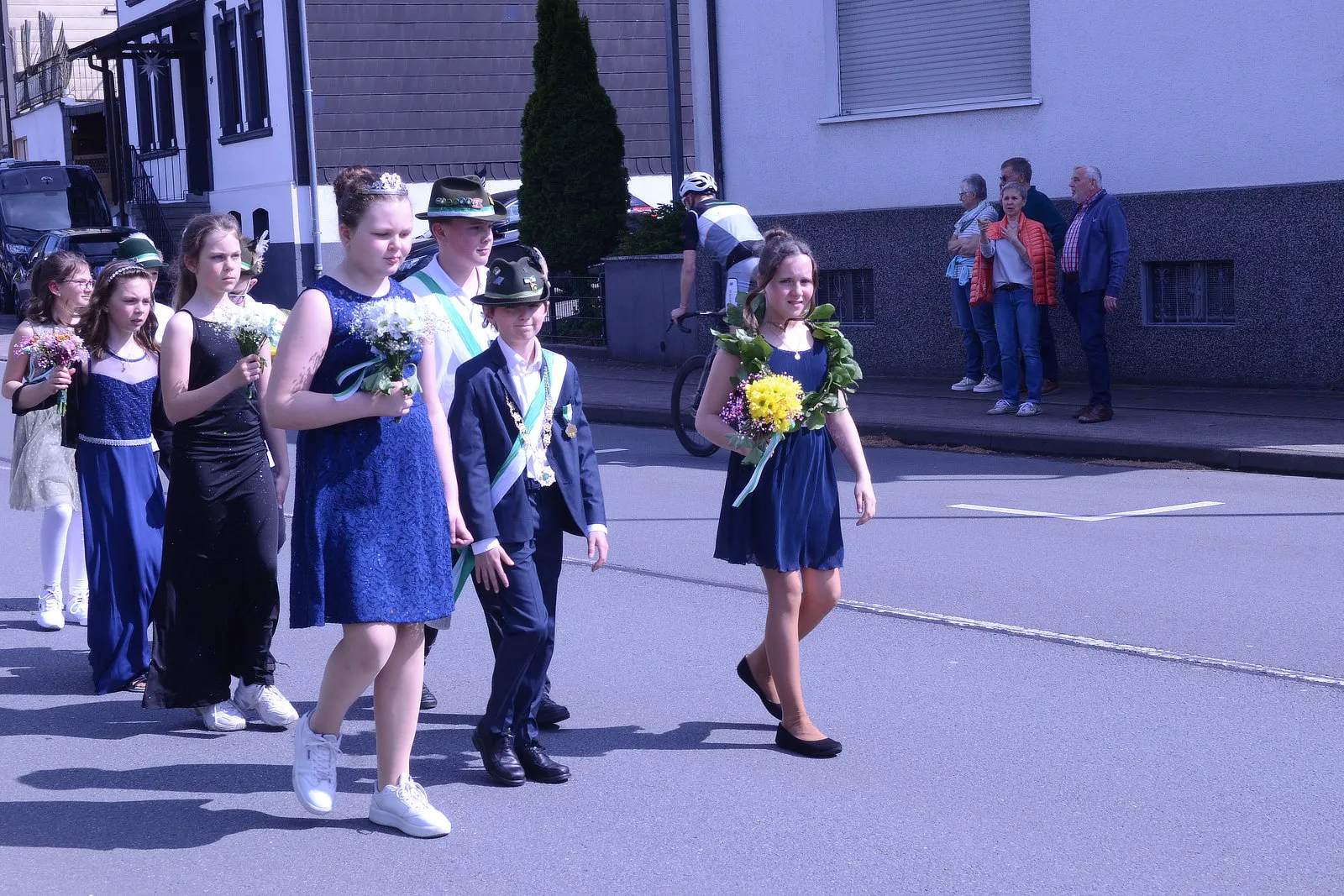 Festumzug mit Jugendlichen in festlicher Kleidung, einige tragen Blumensträuße und Abzeichen. Im Hintergrund stehen Zuschauer am Straßenrand.
