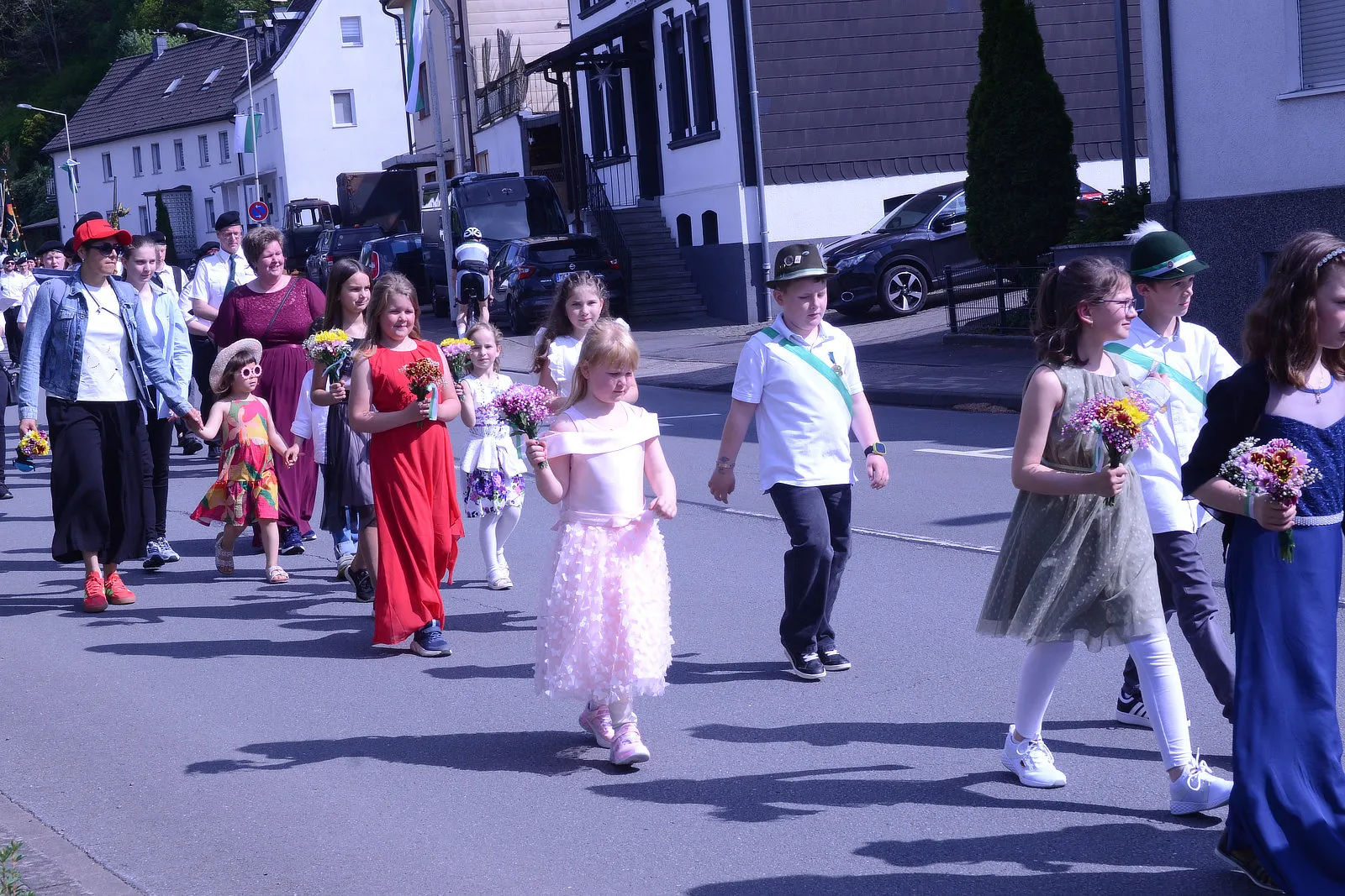 Festumzug mit Kindern und Erwachsenen, die Blumensträuße tragen, vor Häusern und geparkten Autos in Plettenberg.