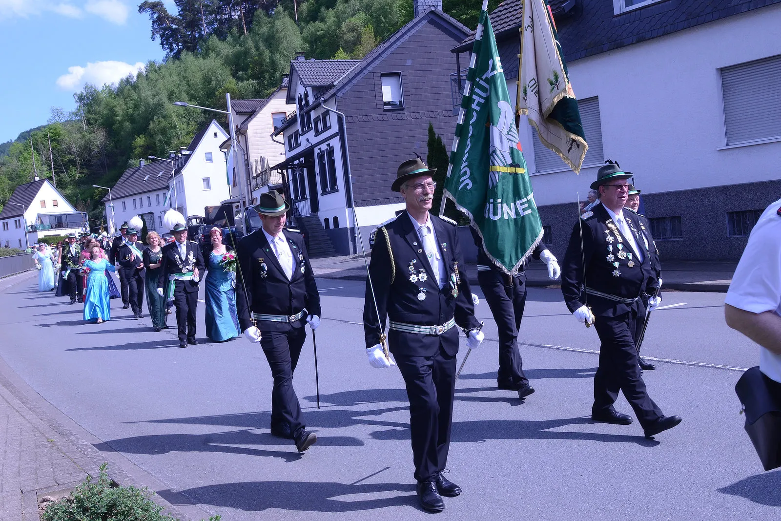 Festzug mit Schützen in historischen Uniformen und einer grün-weißen Fahne durch eine Wohngegend.