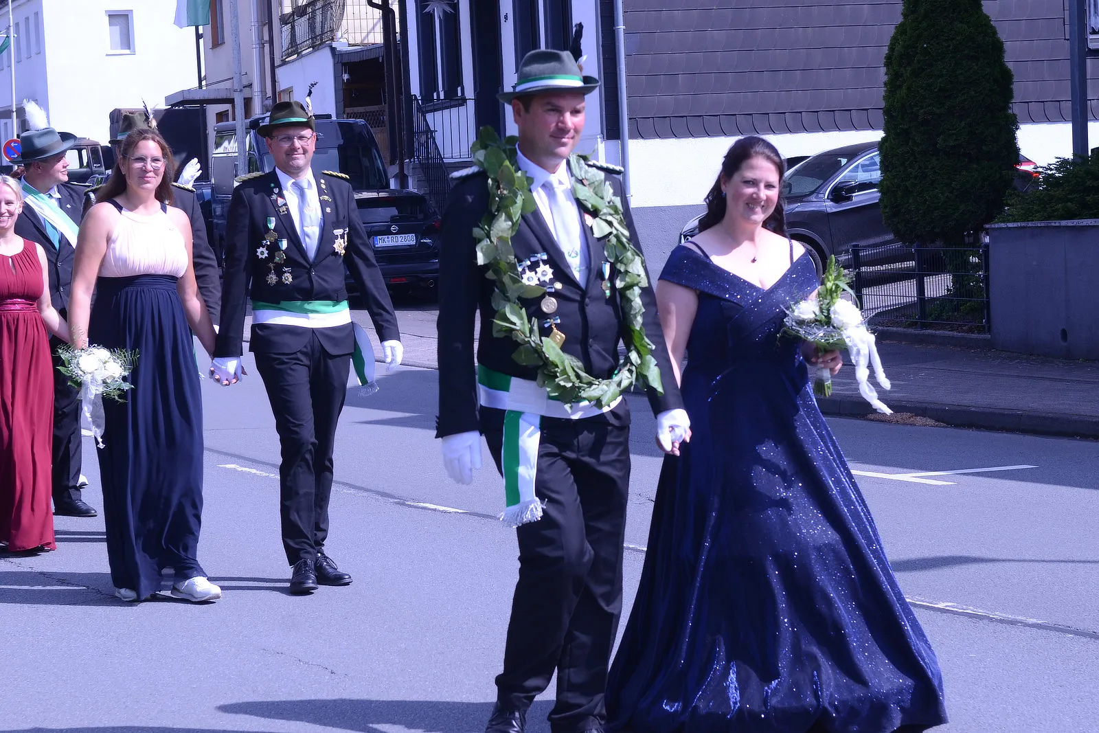 Festzugsteilnehmer in traditioneller Kleidung mit grünen Kränzen und Orden gehen auf einer Straße. Eine Frau trägt ein dunkles Abendkleid, während andere in Anzügen und Kleidern folgen.
