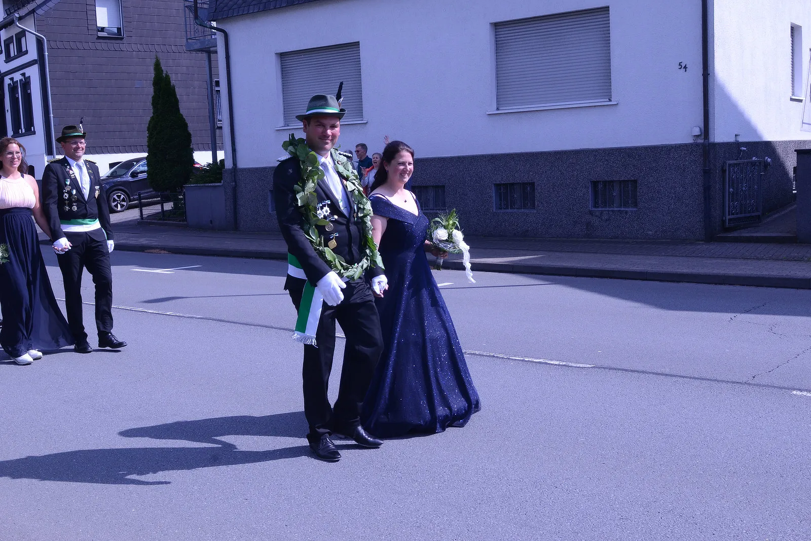 Paar in Festkleidung schreitet während eines Schützenfestumzugs die Straße entlang, der Mann trägt einen grünen Hut und eine Girlande, die Frau ein dunkles Ballkleid und einen Blumenstrauß.