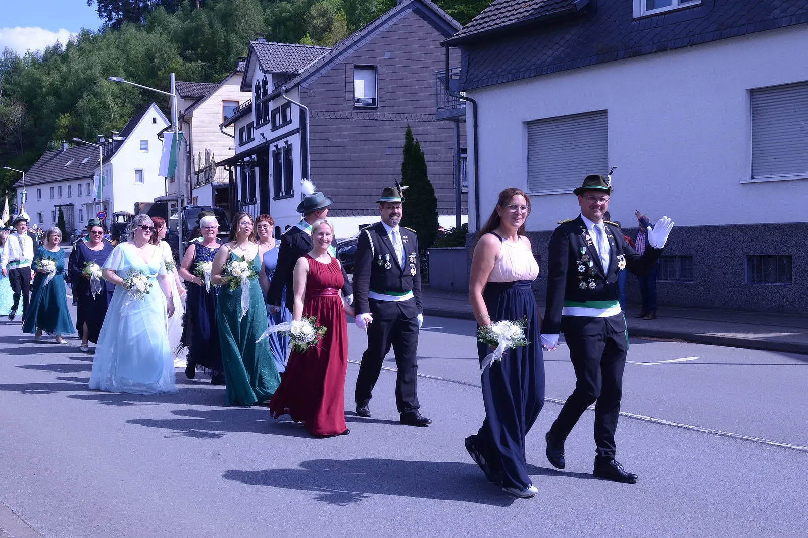 Festumzug mit mehreren Personen in festlicher Kleidung, darunter ein Mann in Uniform und Frauen in Abendkleidern, die Blumensträuße tragen. Im Hintergrund sind Häuser und Bäume zu sehen.