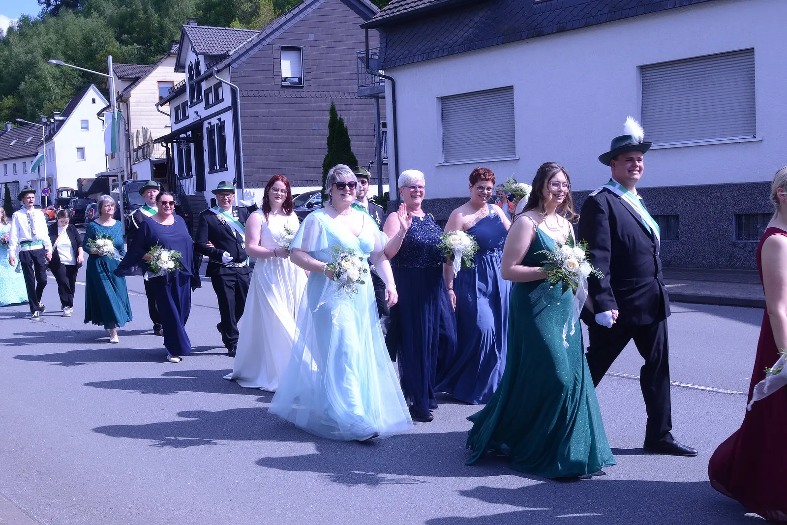Festumzug mit mehreren Personen in festlicher Kleidung, darunter lange Kleider und Anzüge, die Blumensträuße tragen. Im Hintergrund sind Häuser und Bäume zu sehen.