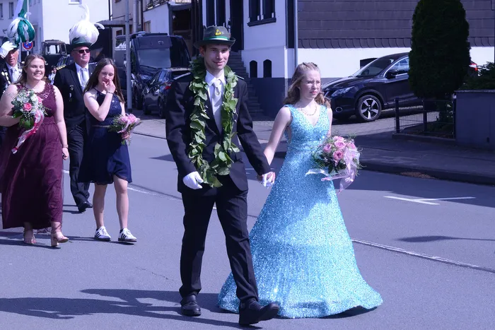 Ein Paar in festlicher Kleidung geht Hand in Hand durch eine Straße, umgeben von weiteren Personen in Tracht und einem Musiker mit einer großen Trompete. Im Hintergrund sind Gebäude und Autos zu sehen.