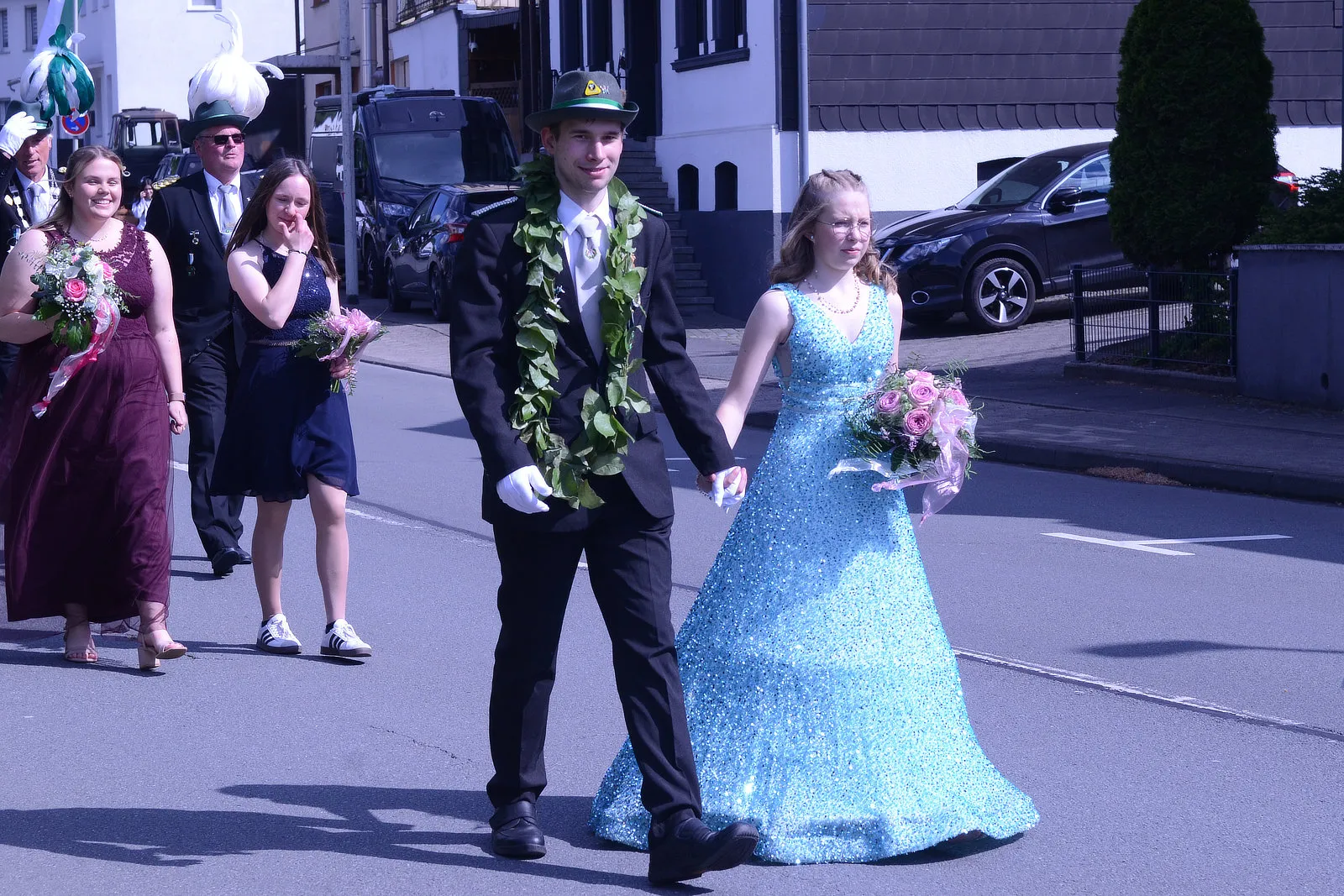 Ein Paar in festlicher Kleidung geht Hand in Hand durch eine Straße, umgeben von weiteren Personen in Tracht und einem Musiker mit einer großen Trompete. Im Hintergrund sind Gebäude und Autos zu sehen.